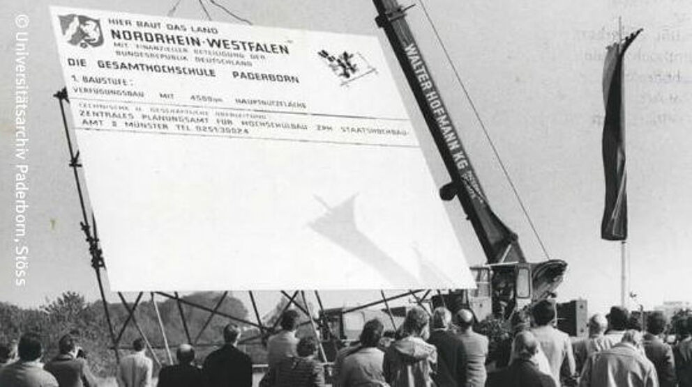 Ein Kran stellt ein großes Schild mit der Aufschrift "Hier baut das Land Nordrhein-Westfalen die Gesamthochschule Paderborn" auf. Die schwarz-weiß Aufnahme zeigt mehrere Zuschauer von hinten.