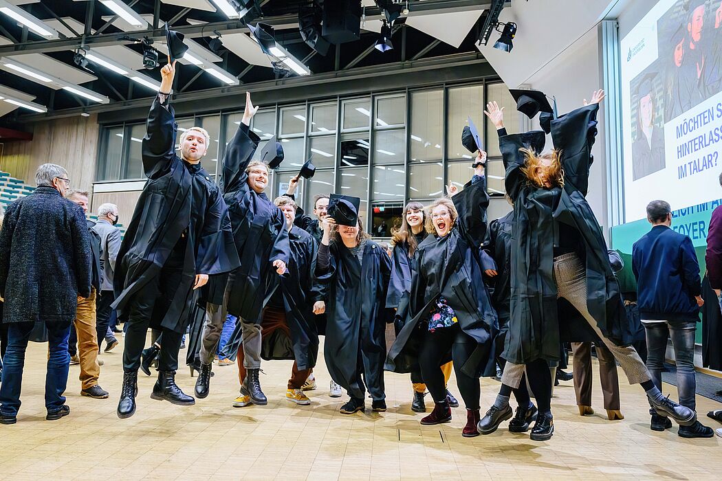 Graduates in the auditorium of Paderborn University wear gowns and throw hats into the air. 