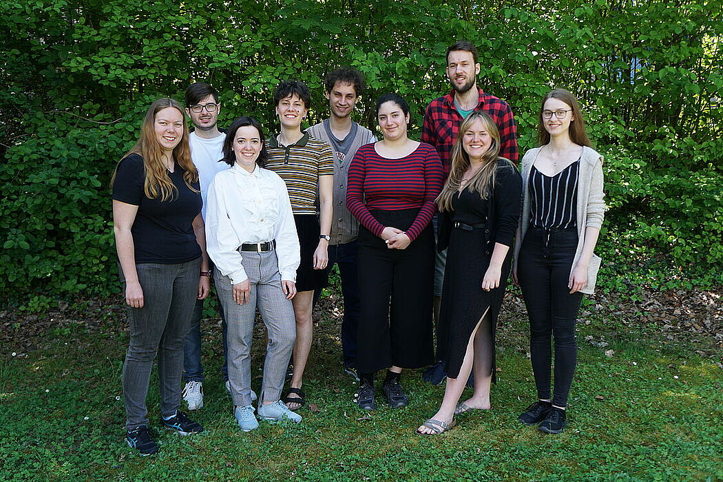 Nine people stand on a meadow in front of green bushes and smile into the camera.