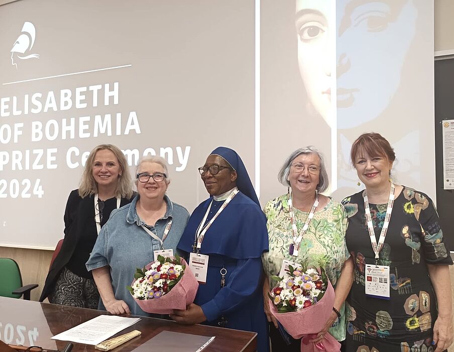 Five women stand next to each other and smile, two of them each holding a bouquet of flowers.