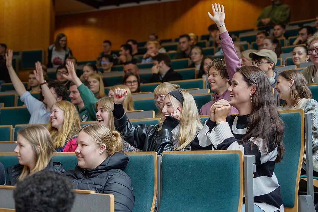 Eine Gruppe von jungen Menschen sitzt im Hörsaal, eine heben die Hand und die Arme und sie lächeln. 