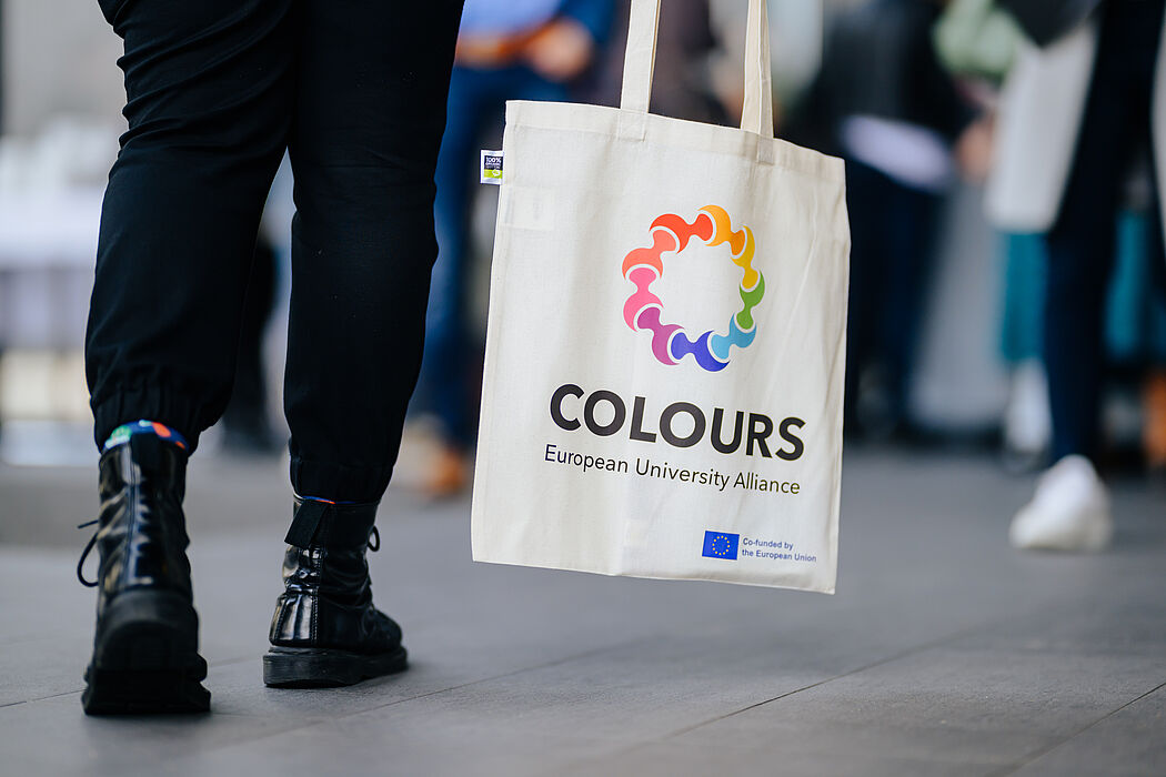 Two legs and a light-coloured bag with the inscription "COLOURS European University Alliance Co-funded by the European Union" and the corresponding logo.