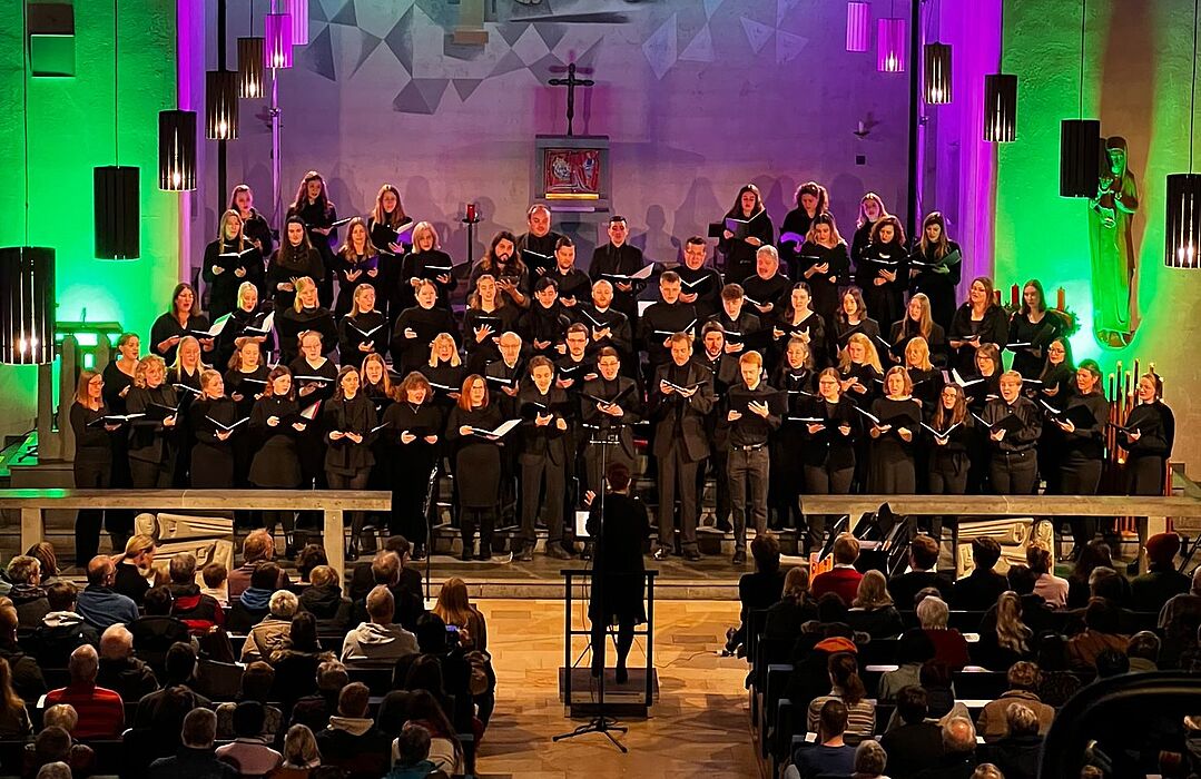 Ein gemischter Chor steht in einer Kirche und singt. Lilafarbenes und grünes Licht sorgt für Akzente.